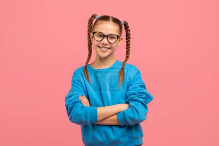 Photo for A cheerful young girl with braided pigtails and eyeglasses stands confidently with arms crossed on a pink background - Royalty Free Image