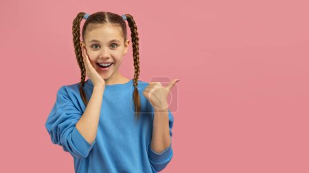 A cheerful young girl with braided hair gestures to her side with a look of happy surprise on a pink background, copy space
