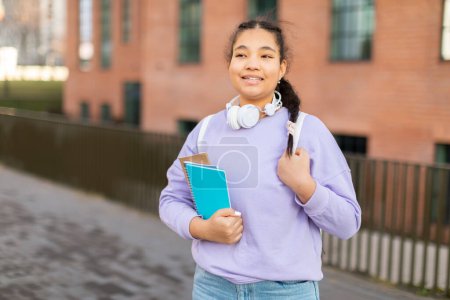 A young girl is standing while holding a book and wearing headphones. Her focus is on the book in her hand, as she seems immersed in reading or listening to music through the headphones.