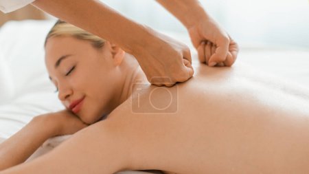 A young woman lies face down, her eyes closed in a serene expression as she enjoys a calming back massage. A professional masseuses hands work skillfully across her back, closeup