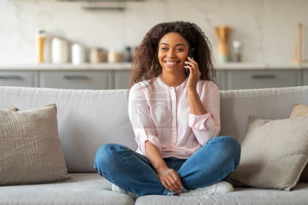 Femme afro-américaine ayant une conversation téléphonique depuis son canapé, représentant la communication moderne à la maison