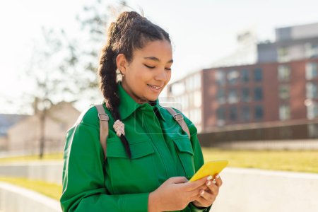 Foto de Una joven con una chaqueta verde vibrante con una mochila sobre su hombro sonríe mientras mira su teléfono inteligente. El sol está emitiendo un cálido resplandor, lo que sugiere que es temprano en el día, posiblemente por la mañana - Imagen libre de derechos