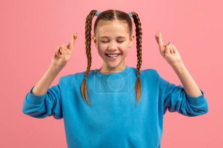 Téléchargez les photos : Une jeune fille joyeuse avec des nattes tressées portant un pull bleu croise ses doigts en espérant bonne chance, sur un fond rose - en image libre de droit