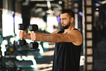 A man is shown holding a pair of dumbbells while standing in a gym. The man appears focused on his workout, showcasing strength and determination in his posture.