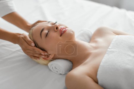 Foto de Una mujer joven se acuesta con los ojos cerrados, totalmente relajada como terapeuta profesional proporciona un masaje relajante en la cabeza, promoviendo el bienestar y el alivio del estrés en un ambiente de spa sereno. - Imagen libre de derechos