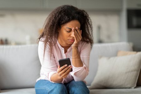 Un momento conmovedor con una mujer afroamericana sentada en casa, apareciendo preocupada mientras revisa su teléfono, evocando empatía