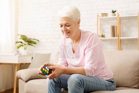 Senior woman is seated on a couch, engrossed in solving a Rubiks Cube with intense concentration. Her hands deftly twist and turn the colorful puzzle