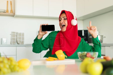 A woman dressed in a vibrant red hijab and green top is captured in a moment of happiness as she sings along to the tunes playing through her white headphones, kitchen interior