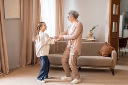 An older woman and a young girl are dancing in a cozy living room. The woman is wearing glasses and a floral dress while the girl is in casual clothes