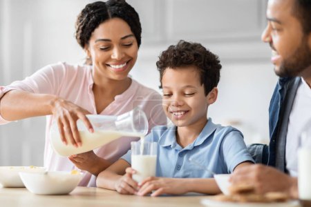 Téléchargez les photos : Une mère afro-américaine verse du lait dans le verre de ses fils pendant que la famille profite d'une routine matinale ensemble dans leur cuisine, embrassant la famille et la santé - en image libre de droit