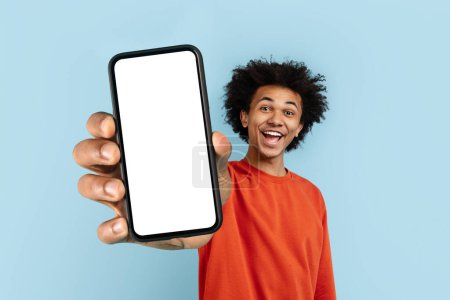 Hombre negro alegre que presenta una pantalla de teléfono móvil en blanco listo para el contenido, aislado en un telón de fondo azul, perfecto para el espacio publicitario