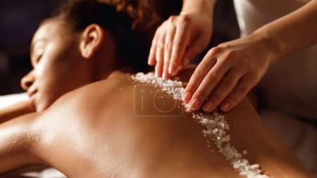 A close-up image of african american woman lying down as she receives a spa salt scrub treatment on her back by a therapist