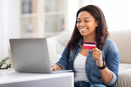 La femme afro-américaine est assise sur un canapé, tenant une carte de crédit et un ordinateur portable. Elle semble être engagée dans des achats en ligne ou des transactions financières, se concentrant intensément sur l'écran de l'ordinateur portable.