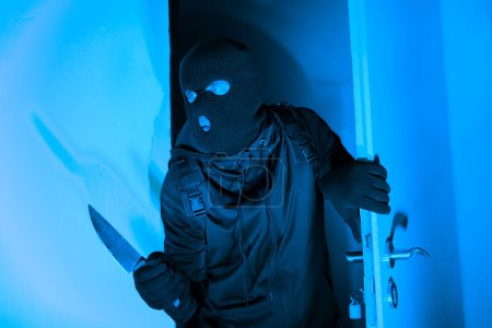 Foto de Un ladrón es capturado en el acto de colarse a través de una puerta con un cuchillo, encarnando la amenaza de un ladrón robando por la noche, evocando miedo en un entorno de apartamento - Imagen libre de derechos