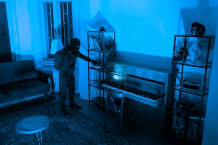 Un ladrón se para junto a un piano en un apartamento, evaluando objetos a robar. El tinte azul transmite una sensación de aislamiento por la noche