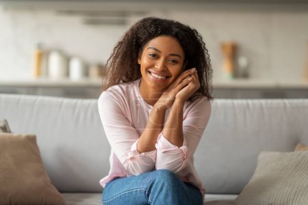 Foto de Joven mujer negra positiva sentada sola en el sofá en casa, tocándose la cara y sonriendo a la cámara. Estilo de vida de los Millennials - Imagen libre de derechos