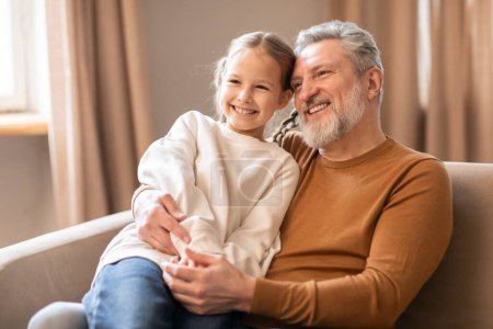 Photo for Elderly man and a little girl are seated on a couch. Grandfather appears to be talking to the girl while she listens attentively. They seem to be engaged in a conversation or storytelling. - Royalty Free Image