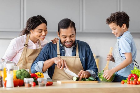 Foto de Alegre familia afroamericana padre madre e hijo se divierten preparando comida en la cocina y riendo juntos - Imagen libre de derechos