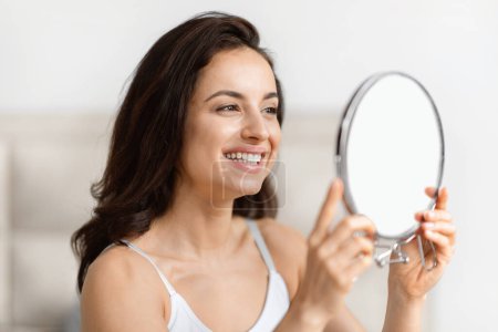 Content millennial brunette pretty lady examines her clear skin, reflecting joy and confidence in a bathroom