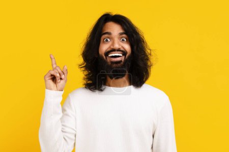 Ein junger indischer Mann mit Bart drückt Begeisterung und Überraschung aus und hebt den Zeigefinger, als würde er eine großartige Idee zeigen. Seine weiten Augen und sein fröhlicher Ausdruck vermitteln ein Gefühl plötzlicher Inspiration
