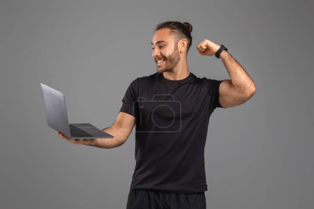 A man is holding a laptop in one hand and flexing his muscles with the other arm. He appears strong and confident, showcasing his physical prowess while multitasking with technology.