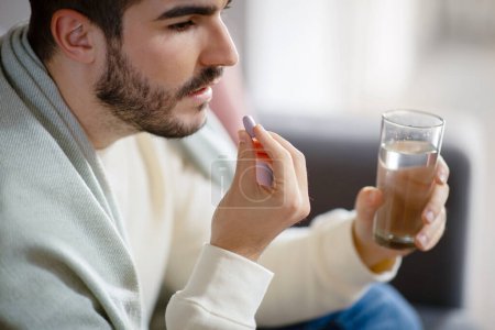 Closeup of focused young man is seated comfortably while holding a pill in one hand and a glass of water in the other, preparing to take his medication