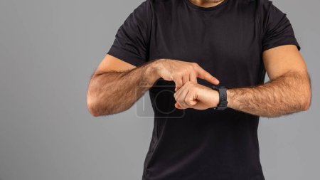 A close-up view of a man in a black t-shirt, his focus on the wristwatch he is checking. The plain gray background emphasizes his action, capturing a moment where time is of the essence, cropped