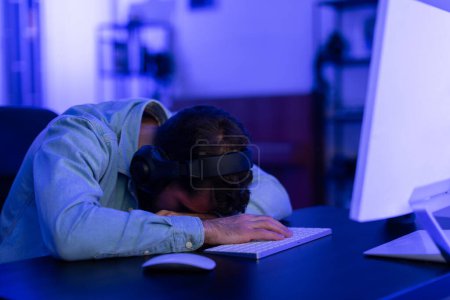 The image conveys a millennial gamer who has fallen asleep from exhaustion at his gaming station at home, suggesting the intense addiction and fatigue that comes with prolonged gaming