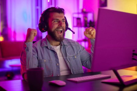 Un hombre milenario muestra un arrebato de felicidad, agarrando sus auriculares de juego, retratando la alegría y la adicción de los juegos en casa