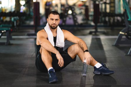 Un hombre con ropa deportiva sentado en el suelo de un gimnasio, rodeado de equipo de ejercicio. Parece estar tomando un descanso o estirándose después de un entrenamiento..