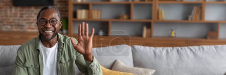 Foto de Imagen de hombre afroamericano de mediana edad sonriente saludando con la mano a la cámara, saluda, dice hola, tiene video llamada en el interior de la sala de estar. Reunión remota, blog moderno, trabajo y webinar en casa, espacio de copia - Imagen libre de derechos