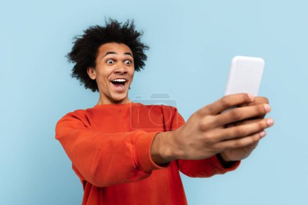 Die Freude eines Schwarzen, als er ein Selfie knipst, isoliert vor einem strahlend blauen Hintergrund, sein Gesichtsausdruck voller Aufregung und Engagement