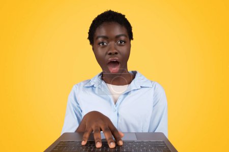 Expressing shock, this african american woman, zoomer of generation z, is surprised at her laptop isolated on yellow