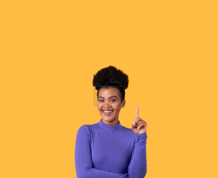 Photo for Hispanic young woman stands against a vibrant yellow backdrop, wearing a long-sleeved purple top. She smiles cheerfully, her eyes sparkling with enthusiasm, as she points upwards with her index finger - Royalty Free Image