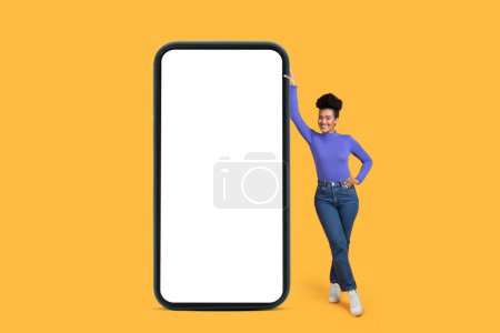 Hispanic woman stands next to a large smartphone with white blank screen isolated on yellow background, mockup copy space