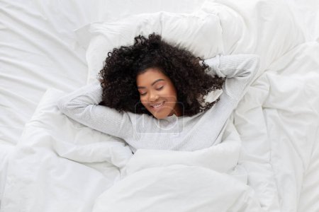 Smiling Hispanic woman is laying down in a bed with crisp white sheets, her eyes closed in relaxation. The room is peaceful and minimalistic, with soft natural light filtering through a window.