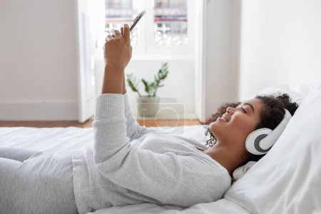 Hispanic woman is laying comfortably on a bed, wearing headphones and listening to music. She appears relaxed and immersed in the sound playing through the headphones.