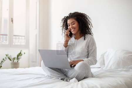 Die hispanische Frau sitzt auf einem Bett und multitasking, indem sie mit ihrem Handy telefoniert, während sie einen Laptop benutzt. Sie wirkt fokussiert und engagiert, balanciert beide Geräte bequem.