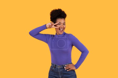 Hispanic woman wearing a purple shirt is smiling. She appears cheerful and content, showing peace gesture next to her eye, yellow background