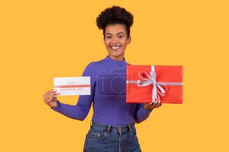 Photo for Hispanic young woman standing while holding two gift cards and a present in her hands, yellow background - Royalty Free Image