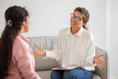 Mujer amigable con gafas y sujetando el portapapeles sentado en el sofá delante del cliente, aconsejando a la joven dama