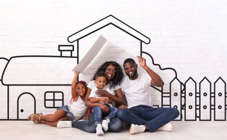 Heureuse famille noire de quatre père mère et les enfants assis sur le sol sur la maison illustrée de leurs rêves sur fond de mur blanc, les parents tenant un toit