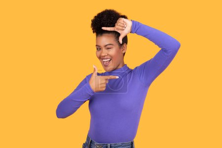 A joyful Hispanic young woman in a purple top is making a frame with her hands, playfully looking through it with a beaming smile.