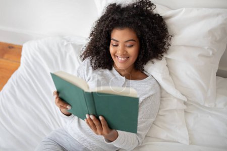 Die hispanische Frau liegt auf einem Bett, in ein Buch vertieft, das sie in ihren Händen hält und lächelt. Der Raum ist schwach beleuchtet, und sie wirkt entspannt und konzentriert auf ihre Lektüre..