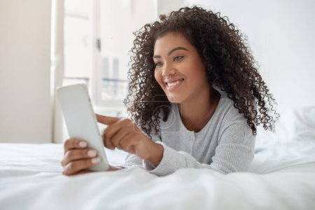Eine fröhliche junge hispanische Frau mit lockigem Haar liegt auf einer weißen Tagesdecke und ist in die Bedienung ihres Smartphones vertieft. Das Tageslicht aus dem Fenster des Zimmers zaubert einen warmen Schein
