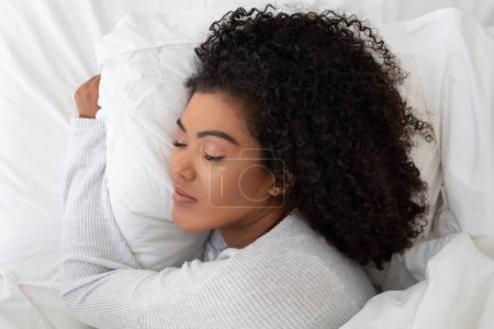Foto de La mujer hispana está recostada en una cama, con la cabeza apoyada en una almohada esponjosa. Ella parece relajada y cómoda, posiblemente preparándose para dormir o tomando un momento para descansar.. - Imagen libre de derechos