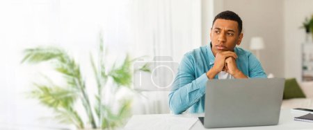 Ein nachdenklicher afrikanisch-amerikanischer Mann, der von zu Hause aus mit Laptop, Stift und Notizblock sowie reichlich Kopierplatz für ein Web-Banner arbeitet