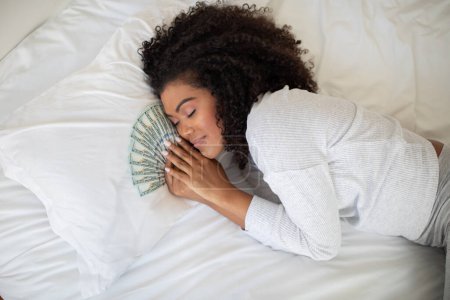 Foto de A la mujer hispana se la ve tumbada en una cama con un fanático del dinero extendido frente a ella. Ella aparece relajada y contenta mientras mira el efectivo en sus manos. - Imagen libre de derechos