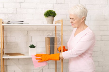 Attractive elderly woman is standing in a room, leaning forward to clean a shelf with a yellow sponge. She is focused on removing dust and grime from the surface.