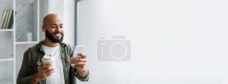 Foto de Hombre negro sonriente disfrutando de un café mientras navega por su teléfono inteligente en una habitación bien iluminada, ideal para banner web con espacio para copiar - Imagen libre de derechos